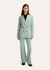 A model stands against a light gray studio background wearing a pale mint green double-breasted suit with wide-leg trousers, paired with a black shirt and black shoes.