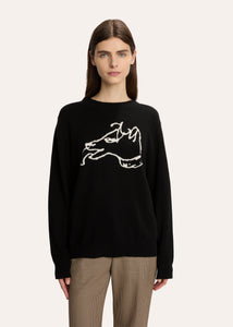 A woman modeling a black crewneck sweater with a white hand-drawn dog graphic on the chest, paired with beige plaid trousers, against a solid off-white background.