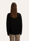 A woman modeling a black crewneck sweater with a white hand-drawn dog graphic on the chest, paired with beige plaid trousers, against a solid off-white background.