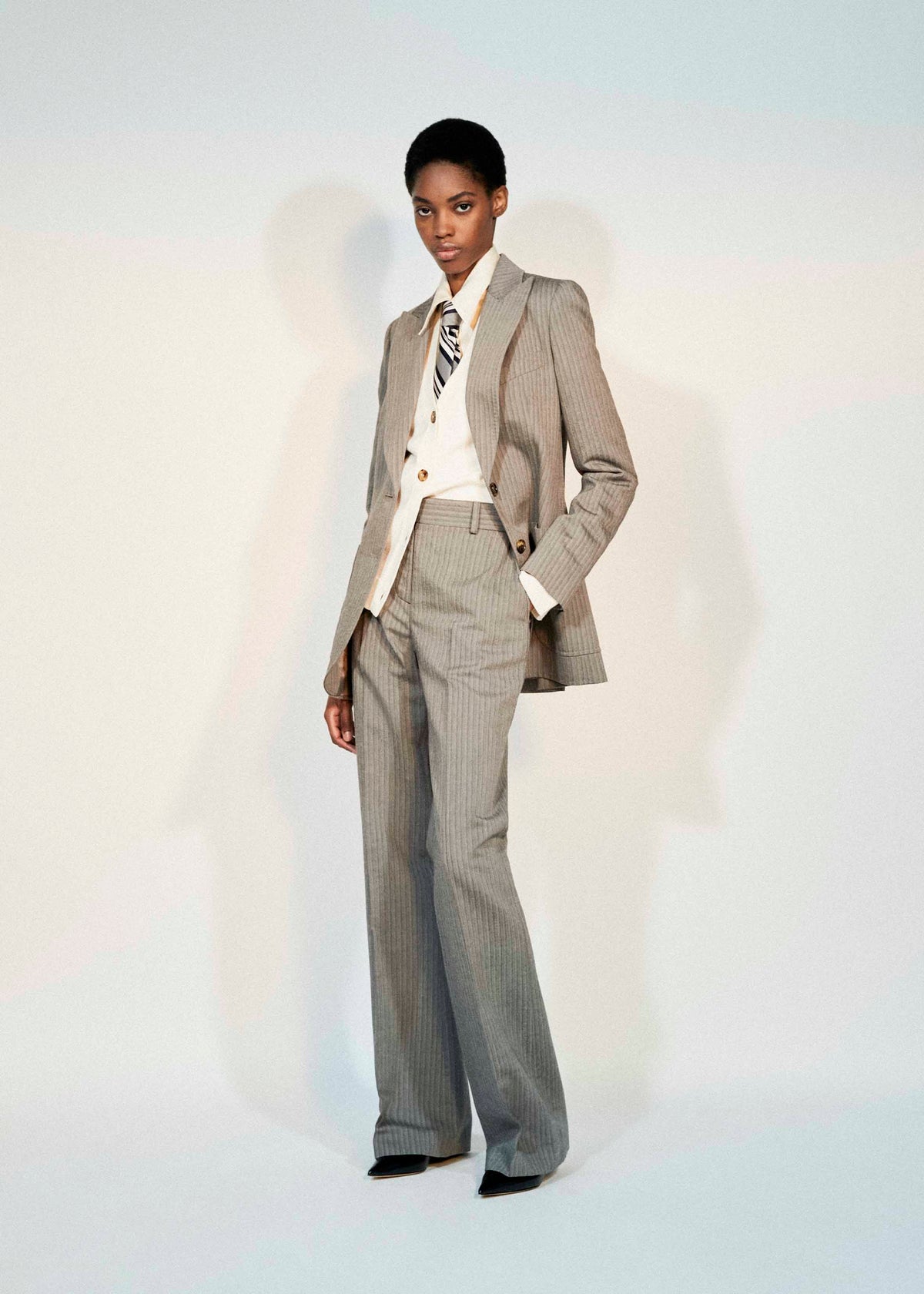 A model stands against a light studio background wearing a tailored light gray pinstripe suit with a white shirt, patterned tie, and black pointed shoes.