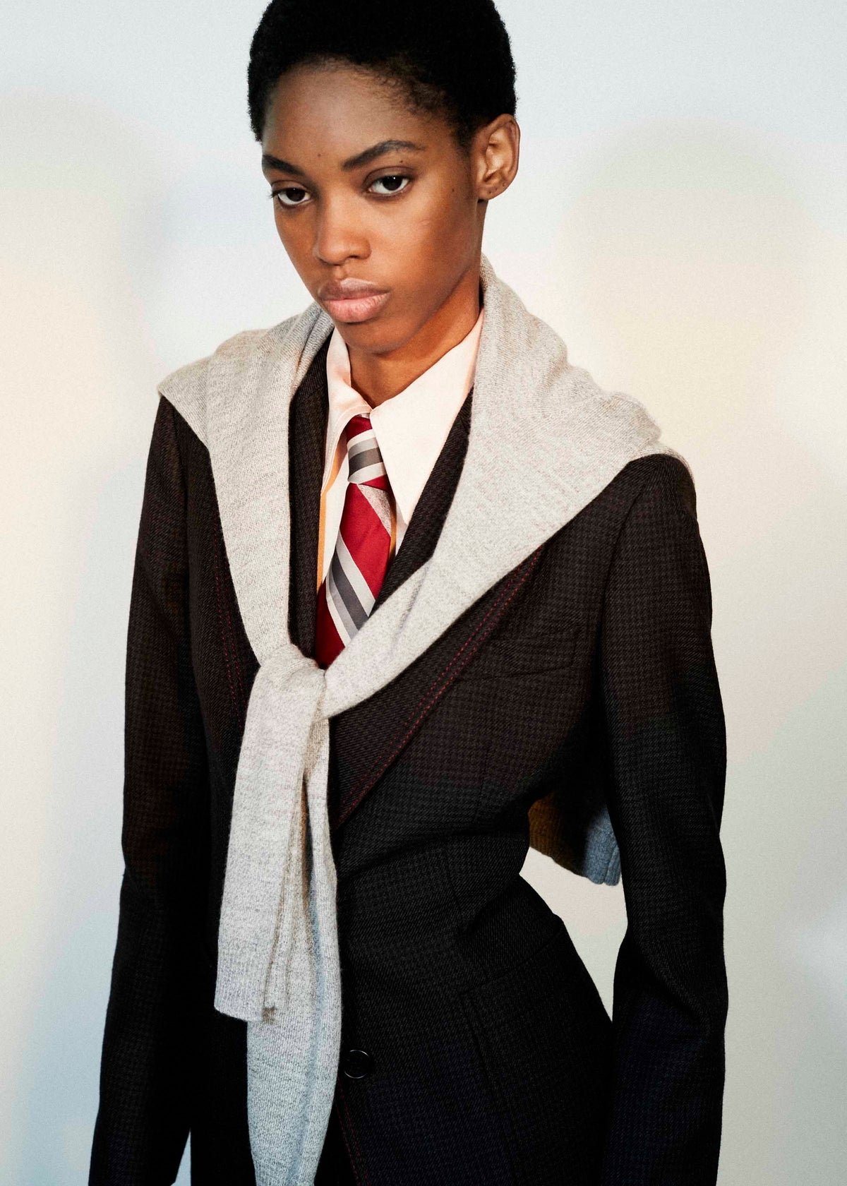 A model wearing a dark brown textured blazer over a white collared shirt with a red and gray striped tie, styled with a light gray knit wrap draped over the shoulders.